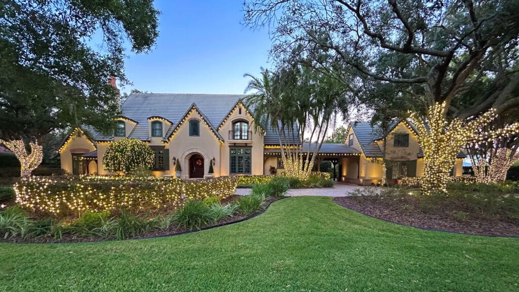 A large, elegant house with steep gabled roofs features a professional Christmas light installation, with warm white string lights illuminating the trees, bushes, and columns, complemented by a neatly manicured lawn and tall front yard trees.