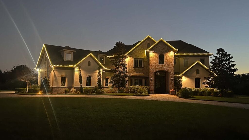 A large house with multiple gables and windows is outlined with warm white string lights at dusk, showcasing a stunning Christmas light installation, surrounded by trees, bushes, and a neatly trimmed lawn.